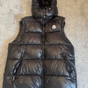 Svart dunväst från Moncler - Säljer en svart, glansig dunväst från Moncler med hög krage och dragkedja framtill. Västen har två fickor med dragkedja och klassisk Moncler-logga på bröstet. Perfekt för lager-på-lager och riktigt snygg till streetwear-stilen.