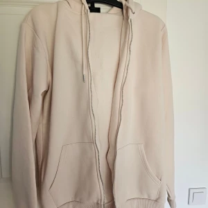 Beige hoodie med dragkedja från SMOG - Ljusbeige hoodie från SMOG i storlek L, slim fit. Tröjan har huva med snörning, dragkedja framtill och två stora fickor. Mjuk bomullskänsla och ribbade muddar vid ärmslut och nederkant. Perfekt för chill dagar eller när du vill ha en clean look.