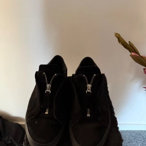 Axel Arigato skor  - Stilrena svarta sneakers från Axel Arigato i mocka med coola silverfärgade dragkedjor framtill istället för klassisk snörning. Skorna har en låg profil och platt sula, perfekt för en clean och modern look. Använda några gånger där av lite billigare pris 