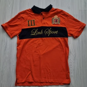 Orange Link Sport pikétröja M - Snygg orange pikétröja från Link Sport med svart krage och svart band över bröstet. Broderad logga och romersk trea på framsidan, samt gul text. Klassisk polo-look med två knappar och korta ärmar. Perfekt för dig som gillar sportig stil.