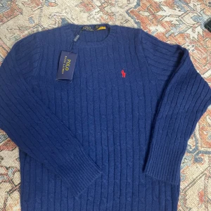 Mörkblå stickad tröja Polo Ralph Lauren - Mörkblå stickad tröja från Polo Ralph Lauren med klassiskt rött broderat logotyp på bröstet. Tröjan har ribbade muddar, rund hals och är långärmad. Perfekt för kyliga dagar och enkel att matcha med jeans eller chinos.