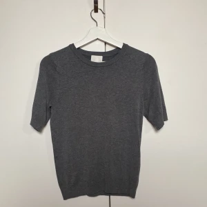 Mörkgrå stickad kortärmad tröja - En stilren mörkgrå stickad tröja med medel korta ärmar och rund halsringning. Tröjan har en enkel och clean look, perfekt för lager-på-lager eller som den är. Mjuk och skön känsla tack vare det stickade materialet. Oanvänd.