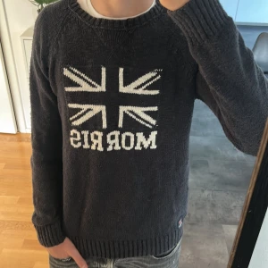 Mörkgrå stickad tröja från Morris - Snygg mörkgrå stickad tröja från Morris med Union Jack-flagga och logga framtill. Tröjan har rund hals, långa ärmar och ribbade muddar. Perfekt för dig som gillar klassisk brittisk stil och vill ha något som sticker ut lite extra. Priset kan såklart diskuteras 