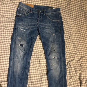 Dondup jeans med slitningar  - Säljer ett par blå jeans från Dondup med coola slitningar och hål på benen. Klassisk femficksmodell med snygga detaljer och kontrastsömmar. Jeansen har en rak passform och är tillverkade i bomullsdenim. Perfekta för dig som gillar en avslappnad och trendig look.
