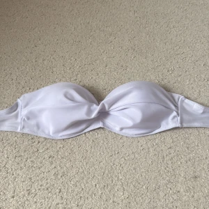 Vit bandeau bikiniöverdel - Snygg vit bandeau bikiniöverdel med twistad detalj framtill. Stilren och enkel design som passar perfekt till stranden eller poolen. Materialet är mjukt och följsamt syntet för skön komfort. Perfekt för dig som gillar minimalistisk stil.