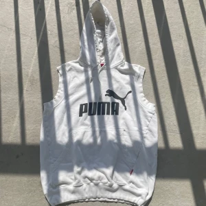 Vit ärmlös hoodie från Puma - Säljer en vit hoodie från Puma med stor svart logga på bröstet. Tröjan är ärmlös, har huva med snörning och en stor magficka framtill. Materialet är mjuk bomull, perfekt för chill dagar eller träning. Snygg och sportig stil som sticker ut.