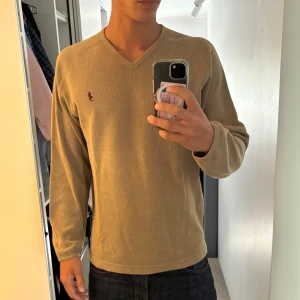 Beige v-ringad tröja Marlboro Classics - Säljer en beige v-ringad tröja från Marlboro Classics i bomull. Tröjan har lång ärm, diskret broderad logga i vinrött på bröstet och ribbade muddar vid hals och ärmslut. Perfekt för dig som gillar enkel och stilren design.