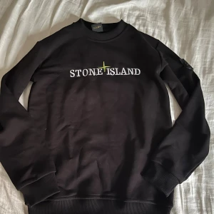 Svart Stone Island sweatshirt - Svart sweatshirt från Stone Island med broderad logga på bröstet och klassisk patch på vänster ärm. Tröjan har rund hals, ribbade muddar och är tillverkad i mjuk bomull. Perfekt för dig som gillar streetwear och vill ha en clean look.