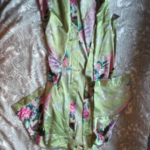 Grön kimono morgonrock Oriental Village - Säljer en lyxig grön kimono-morgonrock från Oriental Village Silk Collection. Klänningen har ett vackert blommigt och fågelmönster i rosa, lila och blå toner, samt knytband i midjan och vida ärmar. Materialet är mjuk silke som känns superfräscht mot huden.