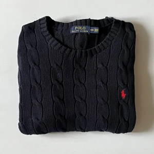 Mörkblå kabelstickad tröja Polo Ralph Lauren - Kabelstickad Mörkblå tröja från Polo Ralph Lauren med klassisk rund hals och röd broderad logga på bröstet. Tröjan är långärmad och har en ribbad kant vid hals, ärmslut och nederkant. Perfekt för dig som gillar stilren och tidlös design.