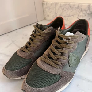 Philippe Model sneakers i grönt och beige - Snygga sneakers från Philippe Model i en mix av grönt och beige mocka och textil. Skorna har olivgröna snören, detaljer i mocka och en vit sula. Insidan har en orange kant och sidan pryds av märkets logga. Perfekta för dig som gillar en sportig och trendig look.