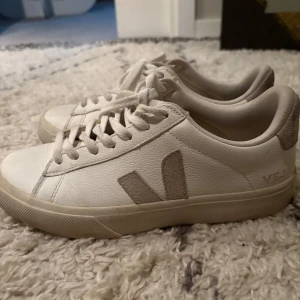 Vita Veja sneakers, strl 38 - Säljer ett par klassiska vita Veja sneakers i skinn med beige mockadetaljer och snörning. Skorna har en platt sula och rund tå, samt den ikoniska V-loggan på sidan.