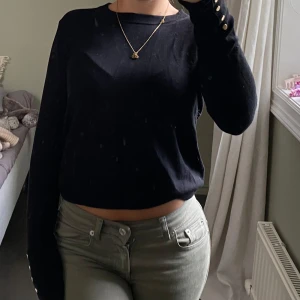 Svart långärmad tröja med knappar - Svart långärmad tröja med rund hals och snygga guldfärgade knappar vid ärmsluten. Tröjan har en enkel och stilren design, perfekt till jeans eller kjol. Materialet är väldigt mjukt och stretchigt, vilket gör den både skön och snygg. Från Zara i XS