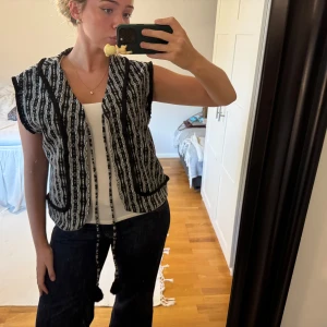 Svartvit mönstrad väst med snörning - Unik svartvit väst med coolt etnoinspirerat mönster och snörning med tofsar framtill. Västen har korta ärmar, två fickor och är i ett mjukt, vävt material. Perfekt att slänga över en enkel topp för en extra touch. Står storlek XL men skulle säga M.