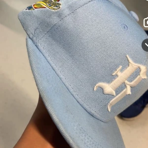 Ljusblå keps med broderad R - Snygg ljusblå keps med vit broderad R på fronten och en färgglad patch på sidan. Klassisk form med böjd skärm och ventilerande hål upptill. Perfekt för dig som vill ha en clean och stilren look med en twist.