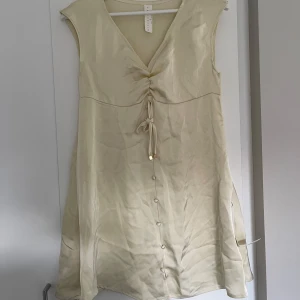 Champagnefärgad klänning Missguided - Supersöt champagnefärgad klänning från Missguided med v-ringning, knytning framtill och dekorativa knappar längs hela framsidan. Ärmlös modell i glansigt material 🤍🤎🤍🤎