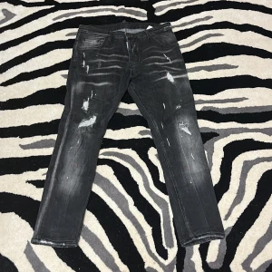 Svarta Dsquared2 jeans med slitningar - Säljer ett par svarta jeans från Dsquared2 med coola slitningar och distressed detaljer. Jeansen har en rak passform och klassisk femficksdesign. Märkespatch bak i midjan. 