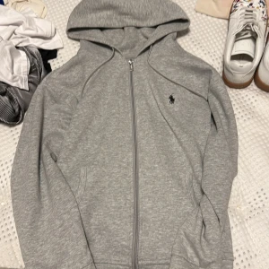  Ralph Lauren hoodie - Snygg grå hoodie från Ralph Lauren med dragkedja och klassisk svart logga på bröstet. Tröjan har huva med snörning, två fickor fram och är tillverkad i mjuk bomullsmix. Perfekt för chill dagar eller när du vill ha en clean och enkel stil. Passar mig bra som är 170