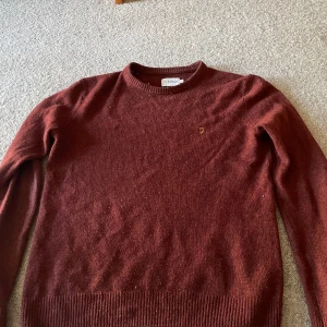 Vinröd stickad tröja från Farah - Mysig vinröd stickad tröja från Farah med rund halsringning och diskret broderad logga på bröstet. Tröjan har långa ärmar och ribbade muddar vid ärmslut och nederkant. Perfekt för kyliga dagar och enkel att matcha med jeans eller chinos.