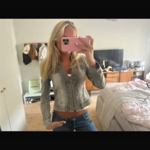 Grå croppad jeansjacka med nitar - Säljer en grå croppad jeansjacka med coola nitar längs knappraden och detaljerade sömmar. Jackan har lång ärm, klassisk krage och knappar framtill. Perfekt för dig som vill ha en edgy och trendig look.