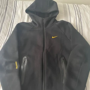 Nocta tracksuit - Svart tracksuit från Nike NOCTA med gul Swoosh och NOCTA-tryck på benet. Byxorna har dragsko i midjan, dragkedjeförsedda fickor och är gjorda i mjukt material som känns riktigt sköna .