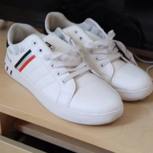 Vita sneakers med färgdetaljer - Snygga vita sneakers med svarta, röda och blå detaljer på sidan och sulan. Skorna har snörning och rund tå, samt svart hälparti. Tillverkade i syntetmaterial med sportig känsla och tryckt text på sidan. Perfekta för dig som gillar stilrena men ändå färgglada skor.