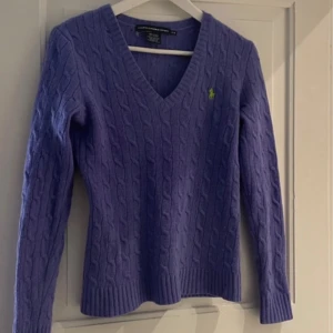 Lila kabelstickad tröja Ralph Lauren - Kabelstickad lila tröja från Ralph Lauren Sport i 100% merinoull. V-ringad modell med lång ärm och klassisk broderad logga på bröstet. Perfekt för dig som gillar en stilren och tidlös look med en touch av färg. Har små hål (se bild) men inget som syns!