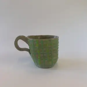 En handgjord kopp i keramik med en brunbeige färg samt gröna rutor. Koppen har ett öra men är även formad för att passa bra i handen. 
