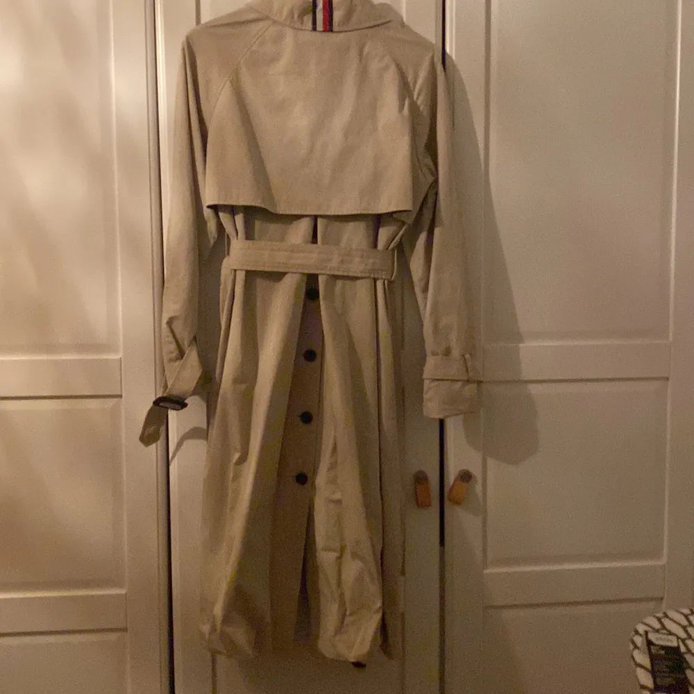 Beige klassisk trench coat från Tommy Hilfiger. Spännen i läder på midjebälte och vid manschetterna. Sprund på baksidan med knappar.. Jackor.