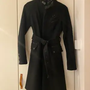 Höst/vinterkappa i svart med dragkedja och skärp. Firgurnära modell.