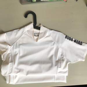 Jättefin vit tight T-shirt i träningdmaterial med liten polokrage. På ena ärmen står det ”budo nord” i svarta bokstäver, och på ryggen finns ett röd/svart kampsportsmärke. Jätteskön och ej genomskinlig💕