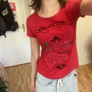 Ed Hardy tröja - Supersnygg röd T Shirt från Ed Hardy. Använt skick, flera ”stenar/paljetter” har lossnat. Står storlek L men sitter som S. Skriv om du har frågor eller vill ha fler bilder. Köparen står för frakt, kan mötas upp