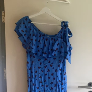 Ganni klänning, storlek 36 - Blå midi klänning med mörkröda blommor från Ganni i storlek 36. Säljer pga jag aldrig använder den, bra i storleken, half shoulder med knut på andra axeln. Superhäftigt tyg
