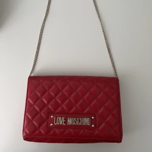 Love moschino i axelbandsmodell, röd  - En axelbandsväska från Love Moschino i färgen röd. Man kan själv välja hur bandet ska sitta och justera det. Slitage förekommer på metallbokstäverna och innerknappen, men inget utöver detta. 