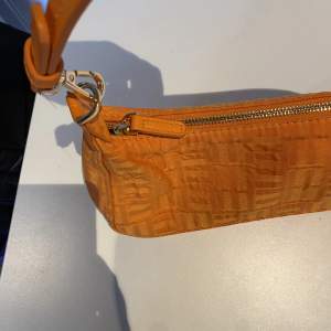 En orange mindre väska (handväska) med en rem, dragkedja samt ett mönster som smälter in som design. Använd fåtal gånger, säljs då den inte kommer till användning hos mig.