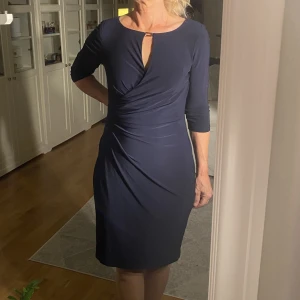 Ralph Lauren klänning  - Marinblå klänning som är draperad i vänstra hörnet vilket ger en figursydd look. Klänningens armar är trekvartslånga. Finns även ett guldfeste vid halsen. 
