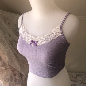 Lila Topp - Söt lila topp med rosett och vit spets. Ett slags mönster me hål i & lite tjockare material så perfekt för en sommardag man inte vill svettas men inte heller frysa:) Använd ett par gånger men väldigt bra skick! Fri frakt fram till 24 Juni. Läs gärna bio<3