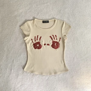 Babytee med tryck - Cool babytee med glittriga röda handavtryck på brösten, från märket Progress! Står ingen storlek men är som en XS/S 🤩