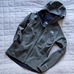 Allväders-jacka The Northface GTX - Olivgrön allväders-jacka / regnjacka för vår och höst i gore-tex. Dammodell: Apex Flex. Stretchigt och tåligt tyg. Strl M. Dragkedja i armhålorna för temperaturreglering. Tre fickor. Bra skick. OBS! Fläck på baksida. Går att tvätta i maskin i 40 grader.