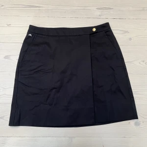 Kjol J.Lindberg  - Svart kort kjol från J.Lindberg. Stängs med knapp och liten hake på insidan. Inneshorts. OBS! Liten reva, se bild nr 2. Annars som i nyskick. Köpt på Erikshjälpen men aldrig kommit till användning.