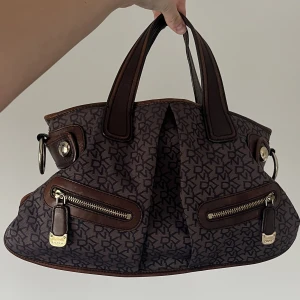 Dkny väska  - Vintage dkny väska.  Tillkommer även ett längre band  Super snygg brun färg tillsammans med de mörklila tyget.  Lite slitningar på baksidan av väskan men överlag jätte bra skick.  För fler bilder och information är de bara att skriva  