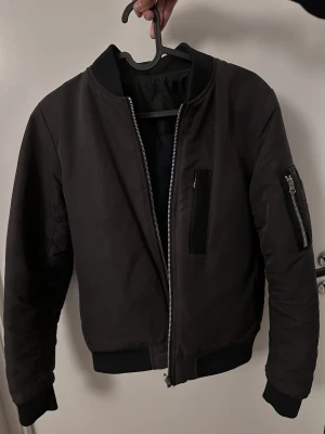 Bomberjacka 2-i-1 - Bomberjacka som kan vändas ut och in, Softshell/vattenavvisande material. Marinblå på insidan.  Svarta sidan har en bröstficka med dragkedja och en liten ficka med dragkedja på ena ärmen. Även 2 vanliga fickor.  Blå sida har 2 vanliga fickor.