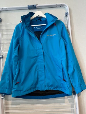 Korallblå regnjacka - Korallblå regnjacka från Everest med röda detaljer. Mycket bra skick. Logga och storlek inuti är lite utsliten.