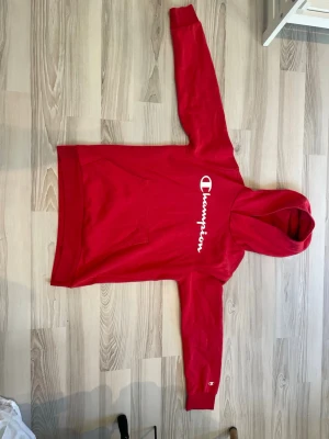 Röd Champion hoodie med huva - Snygg röd hoodie från Champion med stor vit logga på bröstet och klassisk känguruficka framtill. Hoodien har huva och långa ärmar, samt liten Champion-logga på ärmslutet. Perfekt för en avslappnad och sportig stil. Storlek 11/12 år så motsvarar ungefär 146 / 152. 