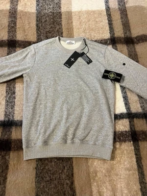 Grå sweatshirt Stone Island, storlek M - Klassisk grå sweatshirt från Stone Island med rund halsringning och ribbade muddar. Tröjan har den ikoniska Stone Island-loggan på vänster ärm och är tillverkad i mjuk bomull. Perfekt för en avslappnad och stilren look.