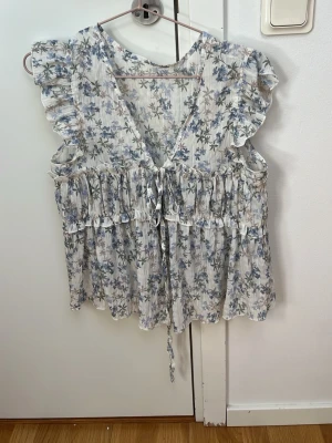 Blommig volangblus med knyt - Supersöt blommig blus i vitt och blått med volangdetaljer och knyt framtill. Blusen har v-ringning, korta volangärmar och ett luftigt, flowy material som passar perfekt till sommaren. Perfekt för dig som gillar romantisk och feminin stil.