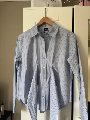 Blåvit randig skjorta med knyte - Prislapp kvar! Snygg blåvit randig skjorta från Gina Tricot med långa ärmar och knytband i midjan. Skjortan har klassisk krage, knappar framtill och breda manschetter. Perfekt för en trendig och avslappnad look.
