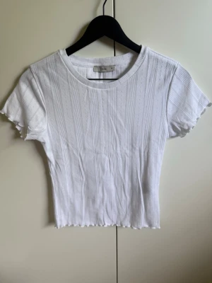 Vit ribbad t-shirt med vågig kant - Säljer en vit ribbad t-shirt med korta ärmar och rund hals. T-shirten har diskreta vertikala ränder och en vågig kant längst ner samt vid ärmsluten. Materialet känns mjukt och stretchigt, perfekt för en avslappnad look. Bara använd några enstaka gånger.