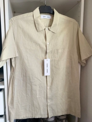 Beige linneskjorta med kort ärm - Snygg beige skjorta i linne med korta ärmar och klassisk krage. Skjortan har knappar framtill och en bröstficka. Perfekt för varma dagar och enkel att styla till olika outfits.