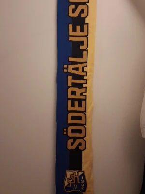 Södertälje sporthalsduk blå och gul - Säljer en lång halsduk i blått och gult med texten 'Södertälje SK' och klubbens logga. Perfekt för att visa ditt stöd på matchen eller hänga upp på väggen. Halsduken har klassisk sportstil och är tillverkad i mjukt material.